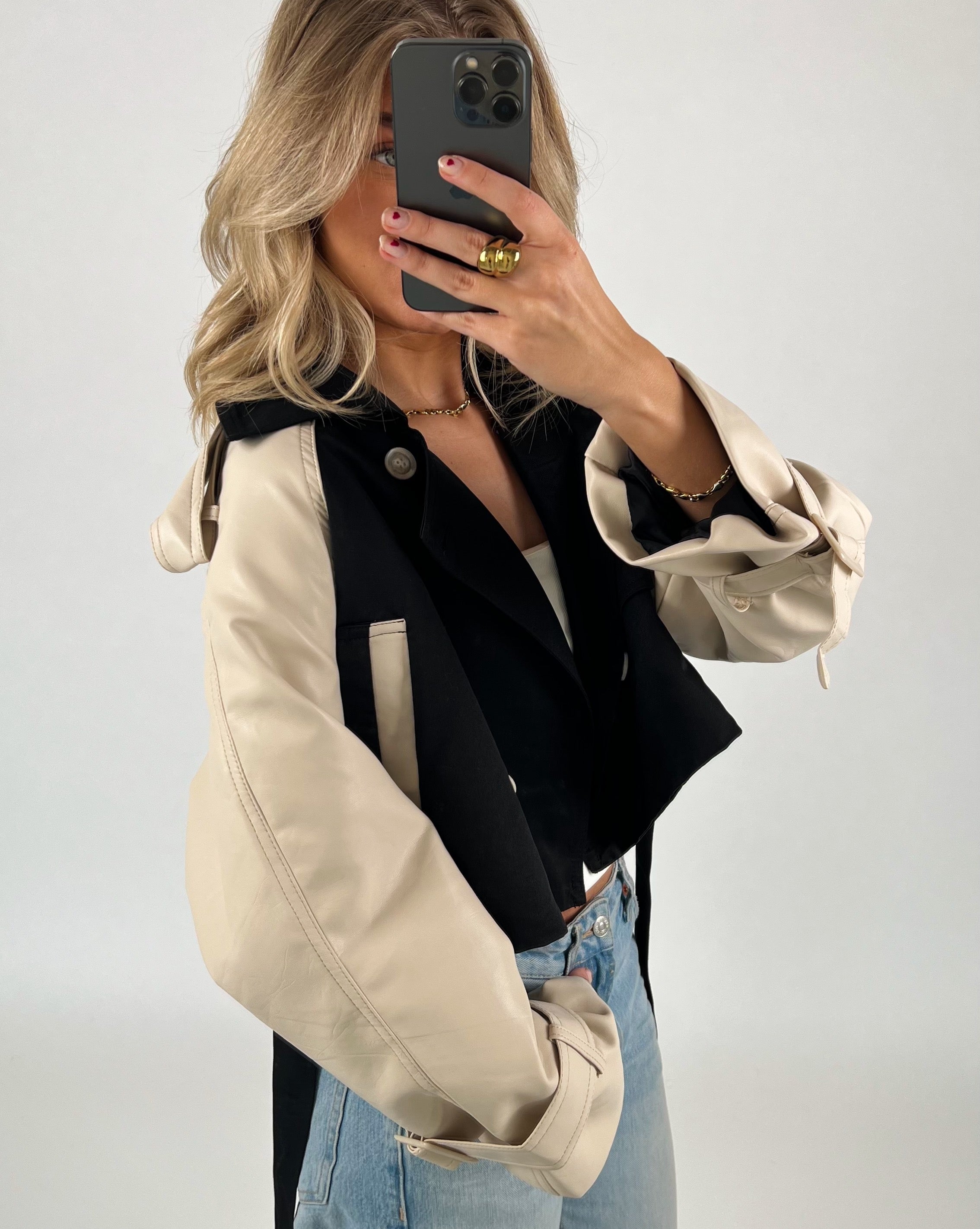 Person tager et spejlbillede iført en sort og beige jakke samt blå jeans. Personen holder en smartphone foran ansigtet i et neutralt baggrundsrum.