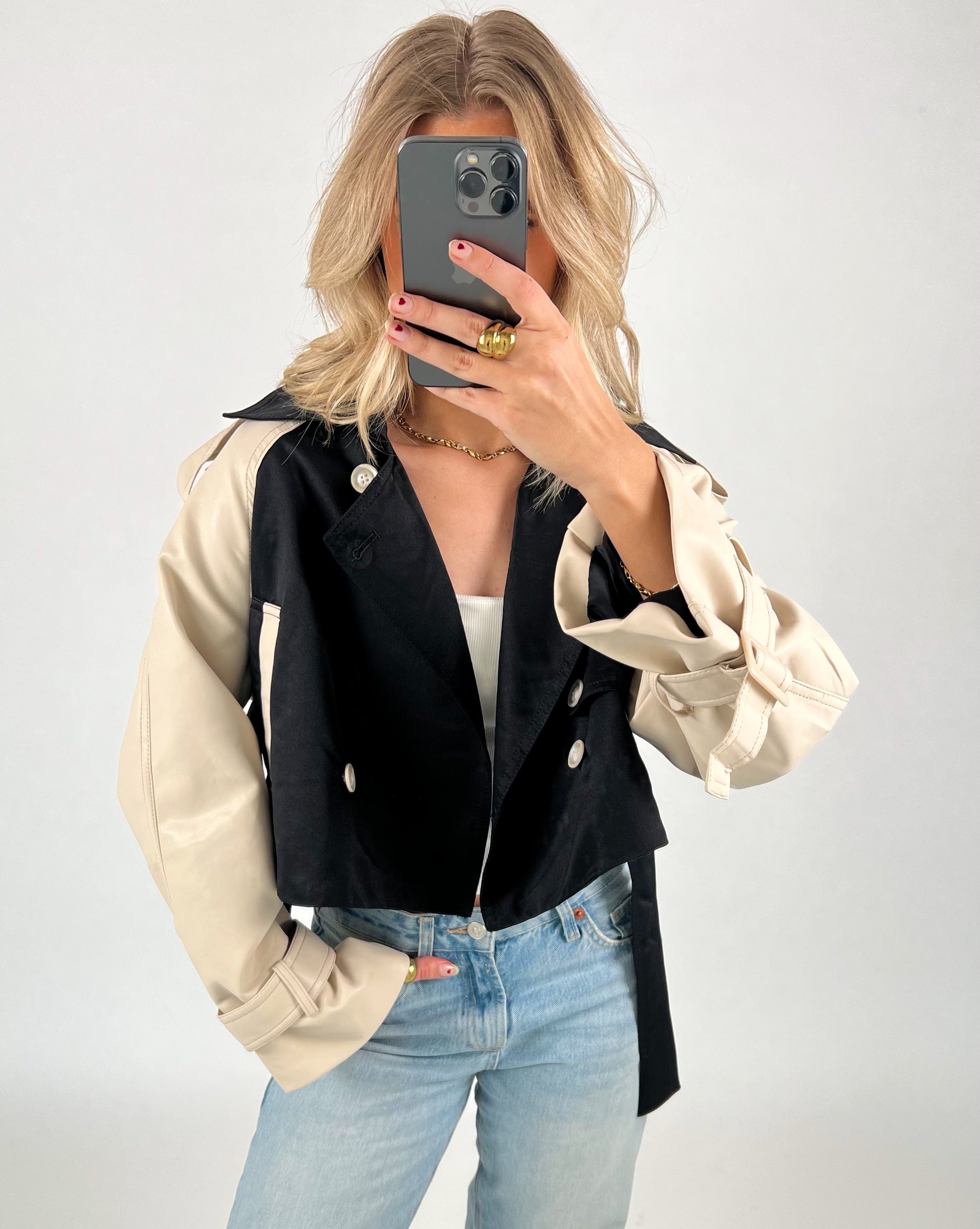 En person tager et spejlbillede iført en sort og beige jakke og blå jeans. Personen holder en telefon op foran ansigtet, omgivet af en lys baggrund.