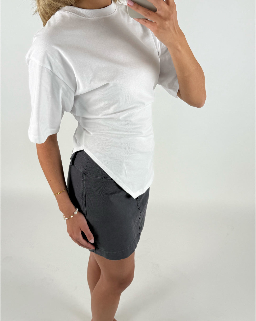 En person iført en hvid t-shirt og mørk nederdel holder en mobiltelefon, fotograferet fra siden i et hvidt studie. Hendes arm bærer to armbånd.