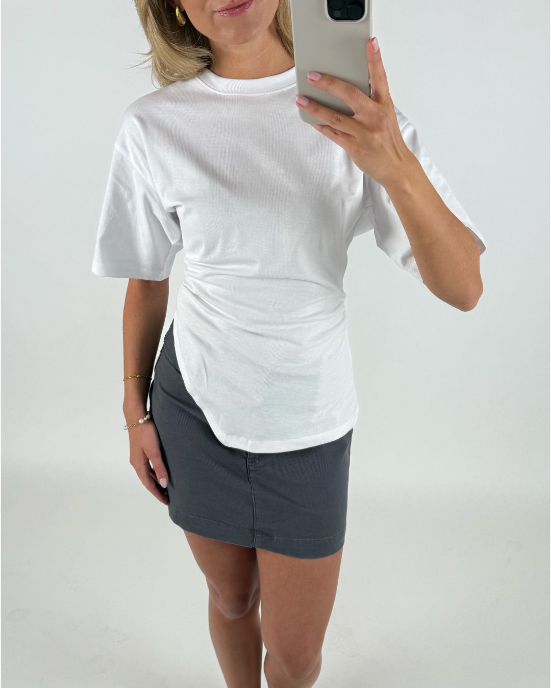 Kvinde iført hvid T-shirt og grå nederdel tager et spejl-selfie. Hun står i et hvidt rum og holder en smartphone med sølvfarvet etui.