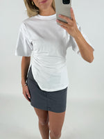 Kvinde iført hvid T-shirt og grå nederdel tager et spejl-selfie. Hun står i et hvidt rum og holder en smartphone med sølvfarvet etui.