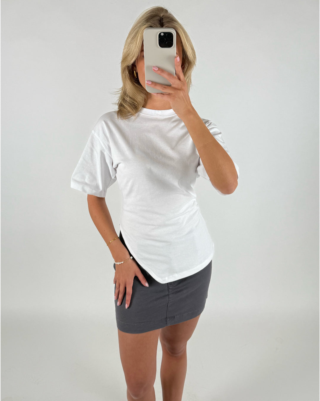 En person iført en hvid t-shirt og en mørkegrå nederdel holder en mobiltelefon foran ansigtet i et minimalistisk, lyst rum.