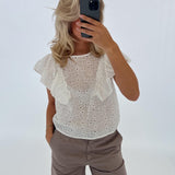 En person i en hvid bluse tager et spejlbillede med en smartphone. Personen står i et lyst rum og har hånden i bukselommen.