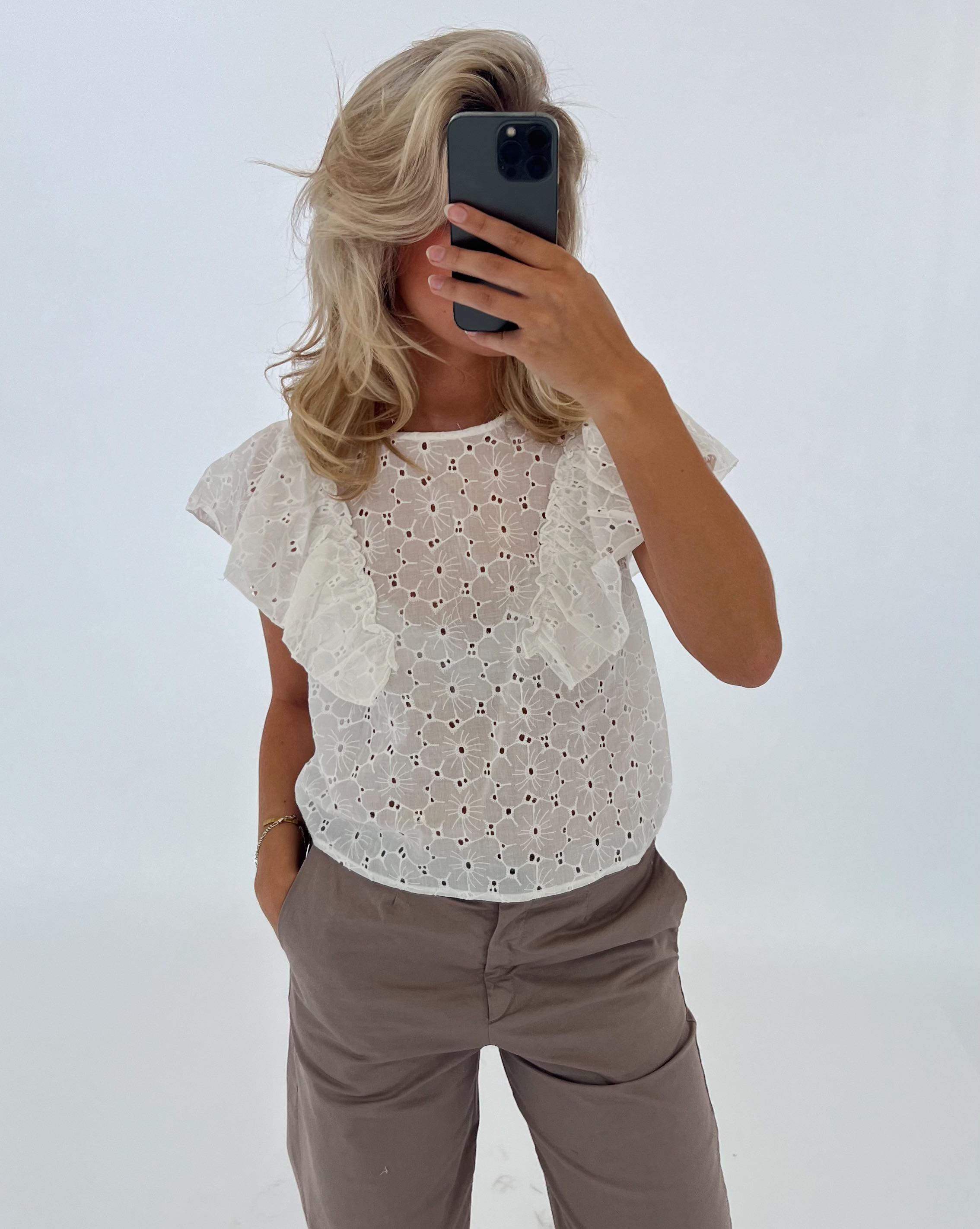 En person i en hvid bluse tager et spejlbillede med en smartphone. Personen står i et lyst rum og har hånden i bukselommen.