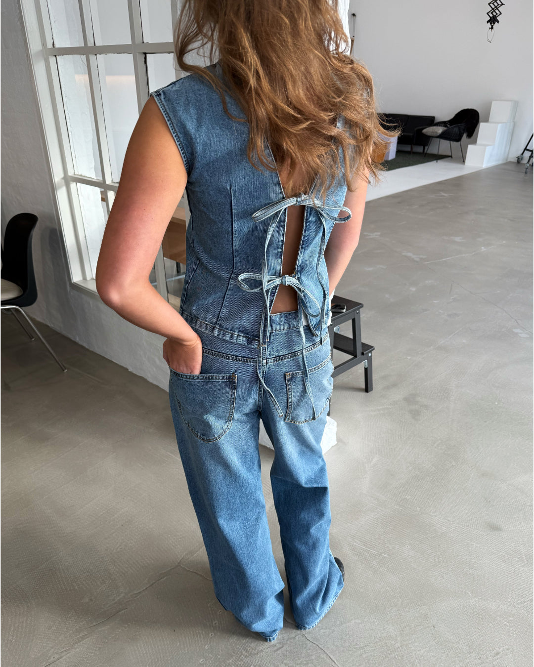 Kvinde iført denimdragt med åbninger og bindebånd på ryggen står i industrielt rum med lyse vægge, store vinduer og moderne møbler.
