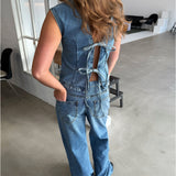 Kvinde iført denimdragt med åbninger og bindebånd på ryggen står i industrielt rum med lyse vægge, store vinduer og moderne møbler.