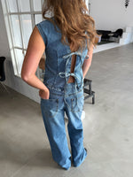 Kvinde iført denimdragt med åbninger og bindebånd på ryggen står i industrielt rum med lyse vægge, store vinduer og moderne møbler.