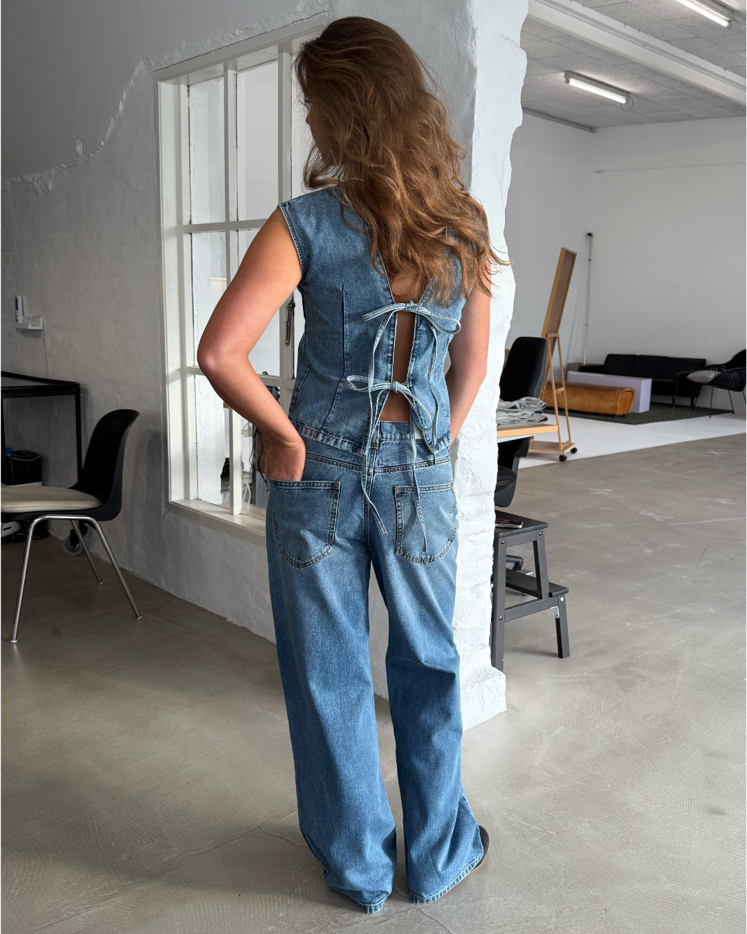 En person iført denim-outfit står med ryggen til og hænderne i lommerne. Lyse omgivelser med moderne møbler i et minimalistisk indrettet rum.