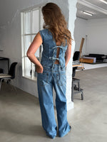En person iført denim-outfit står med ryggen til og hænderne i lommerne. Lyse omgivelser med moderne møbler i et minimalistisk indrettet rum.