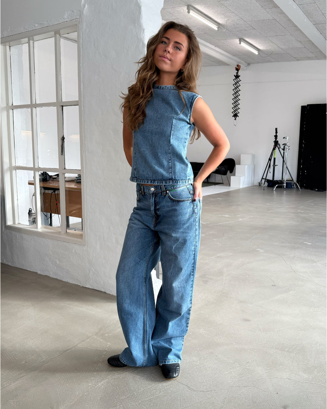 En person iført denim-top og jeans står afslappet i et lyst rum med beton gulv og studieudstyr i baggrunden. En glasvæg viser et tilstødende kontor.