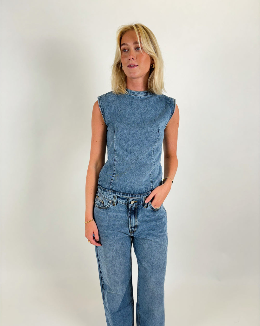 En person iført denimdragt står afslappet mod en lys baggrund. Personen har blondt hår og armene let bøjet ved albuerne, med hænderne tæt på lommerne.