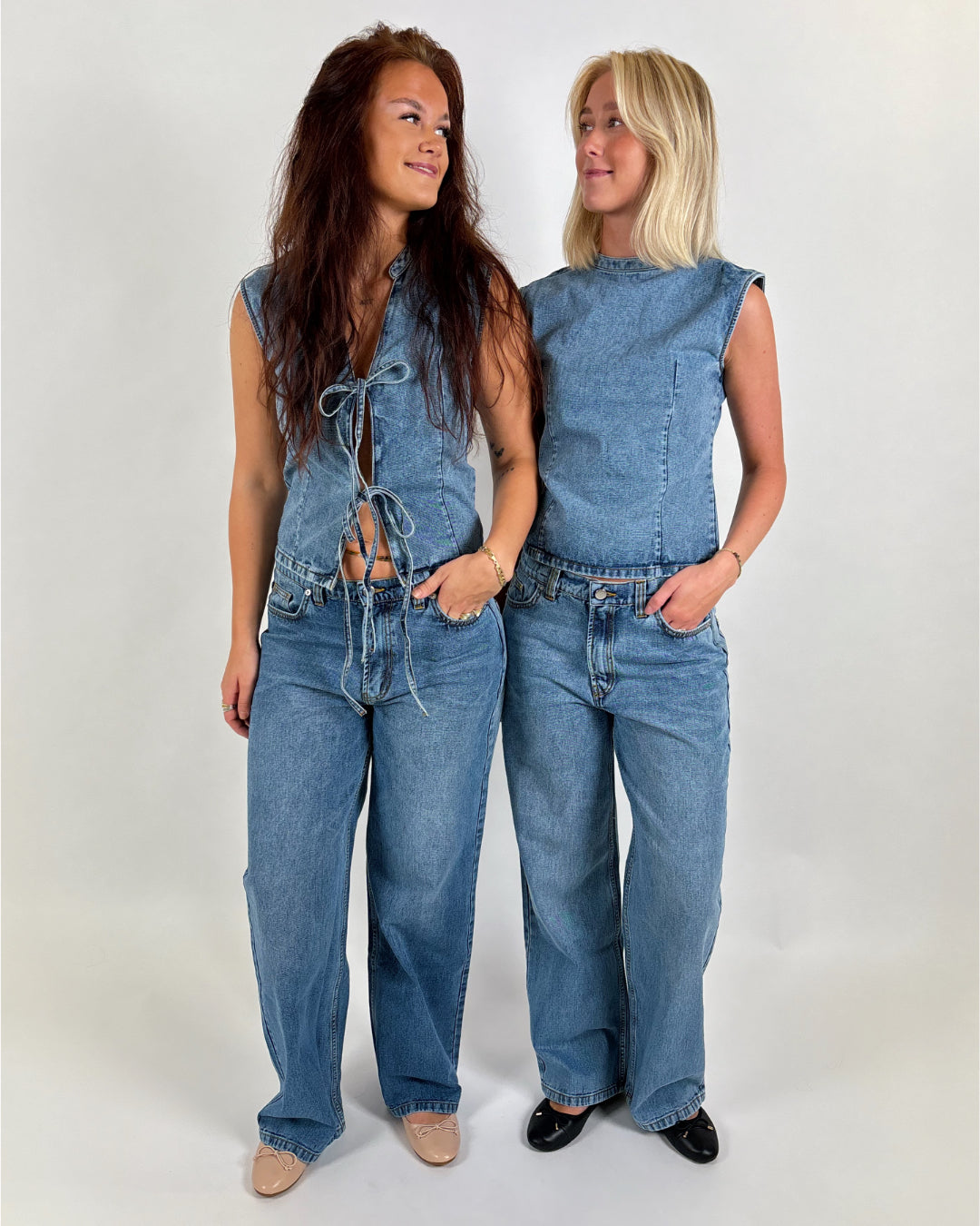 To kvinder iført denim toppe og jeans står smilende og kigger på hinanden i et neutralt rum. Den ene har en blonde-top uden ærmer, den anden en ærmeløs bluse.
