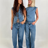 To kvinder iført denim toppe og jeans står smilende og kigger på hinanden i et neutralt rum. Den ene har en blonde-top uden ærmer, den anden en ærmeløs bluse.