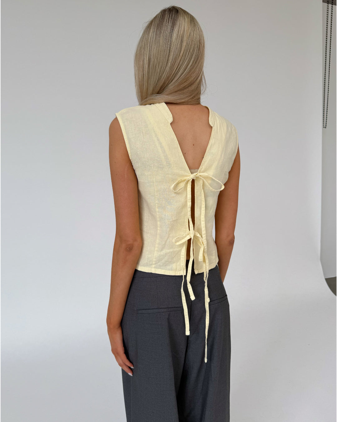 En kvinde med langt blond hår bærer en gul bluse med åbent rygdesign bundet med bånd, står stille i et minimalistisk rum med en rolig baggrund.