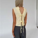 En kvinde med langt blond hår bærer en gul bluse med åbent rygdesign bundet med bånd, står stille i et minimalistisk rum med en rolig baggrund.