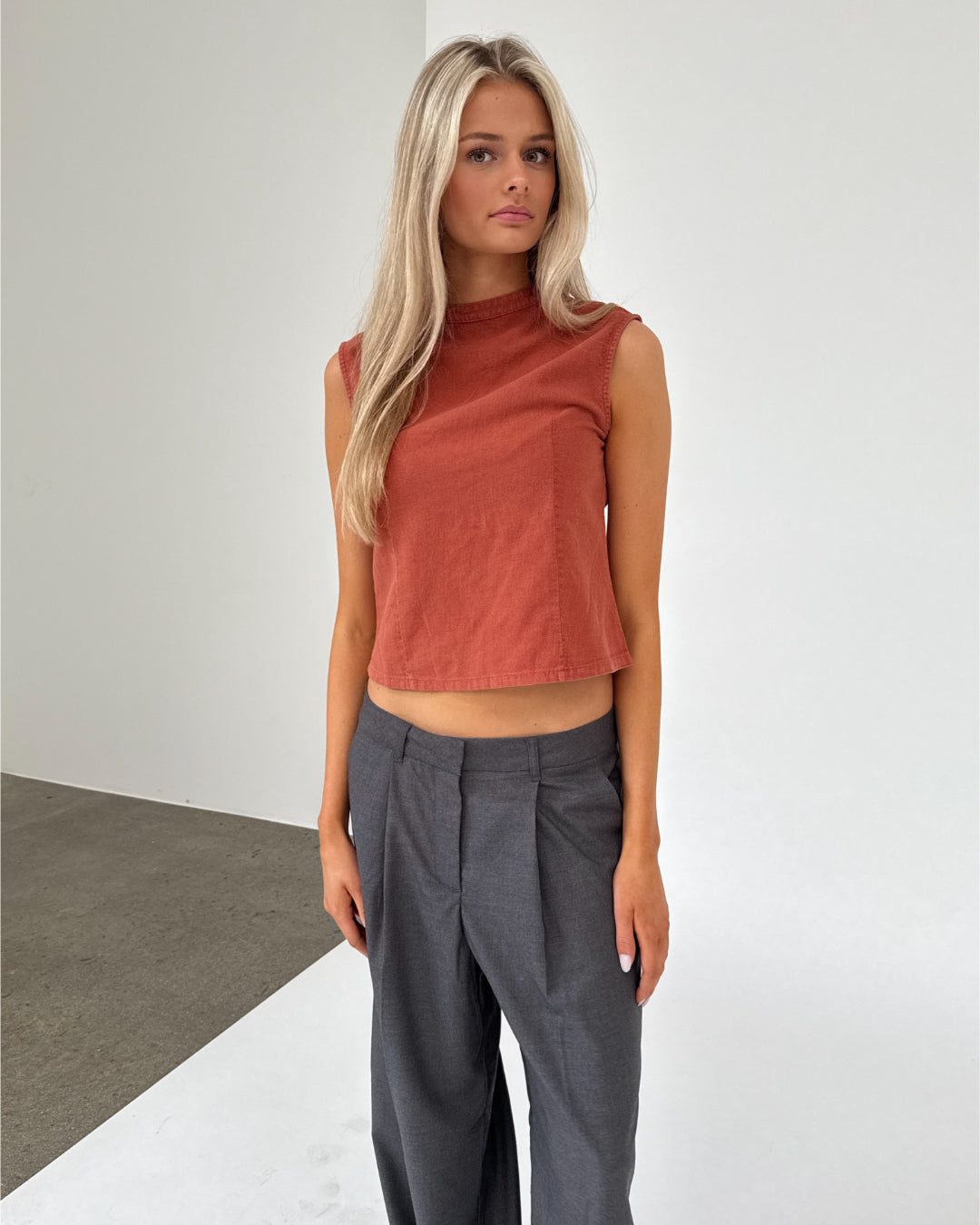 En kvinde står stille iført en ærmeløs orange top og grå bukser. Hun befinder sig i et minimalistisk rum med lyse vægge og mørkt gulv.