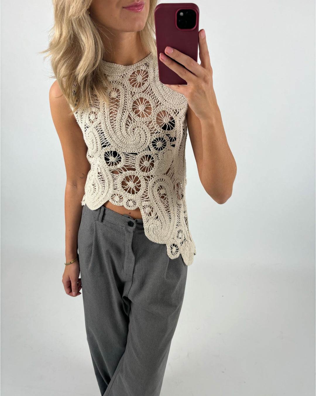 En person tager en selfie i spejlet iført en hæklede beige top og grå bukser. Håret er lyst og krøllet. Baggrunden er simpel og hvid.
