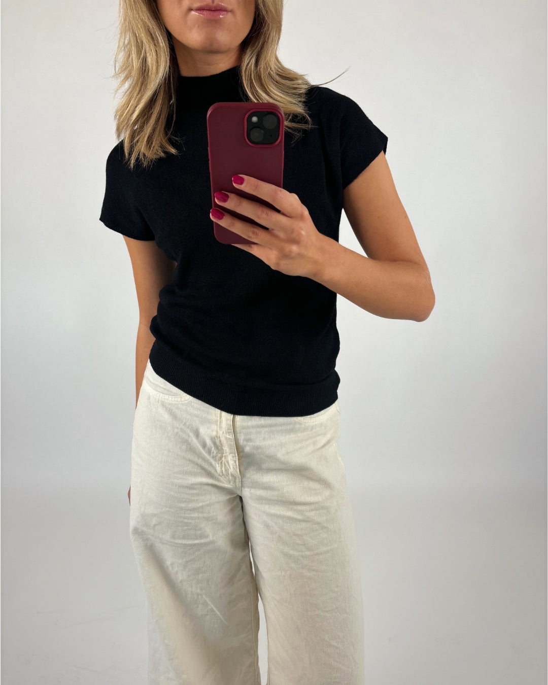 Person holder en mobiltelefon i hånden, taget som selfie. Personen bærer en sort trøje og hvide bukser. Baggrunden er en lys væg.