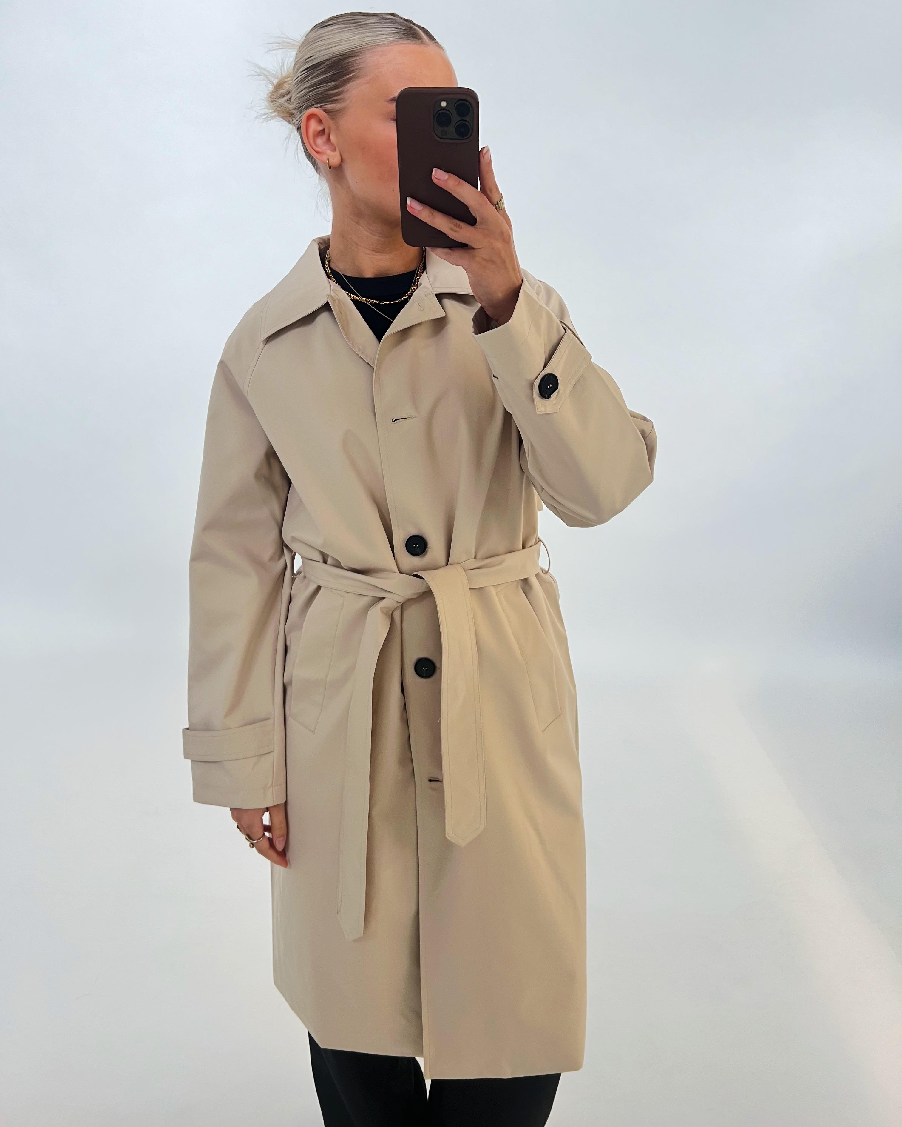 En person iført en beige trenchcoat holder en smartphone foran ansigtet. Baggrunden er ensfarvet hvid, hvilket fremhæver jakken og telefonen.