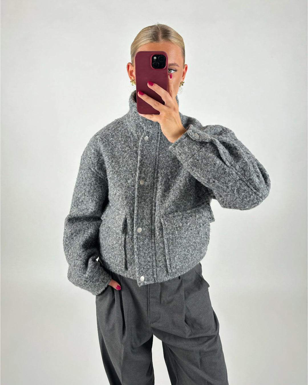 En person iført en grå uldjakke tager et spejlbillede med en smartphone. Personen står i et minimalistisk rum med en neutral baggrund.