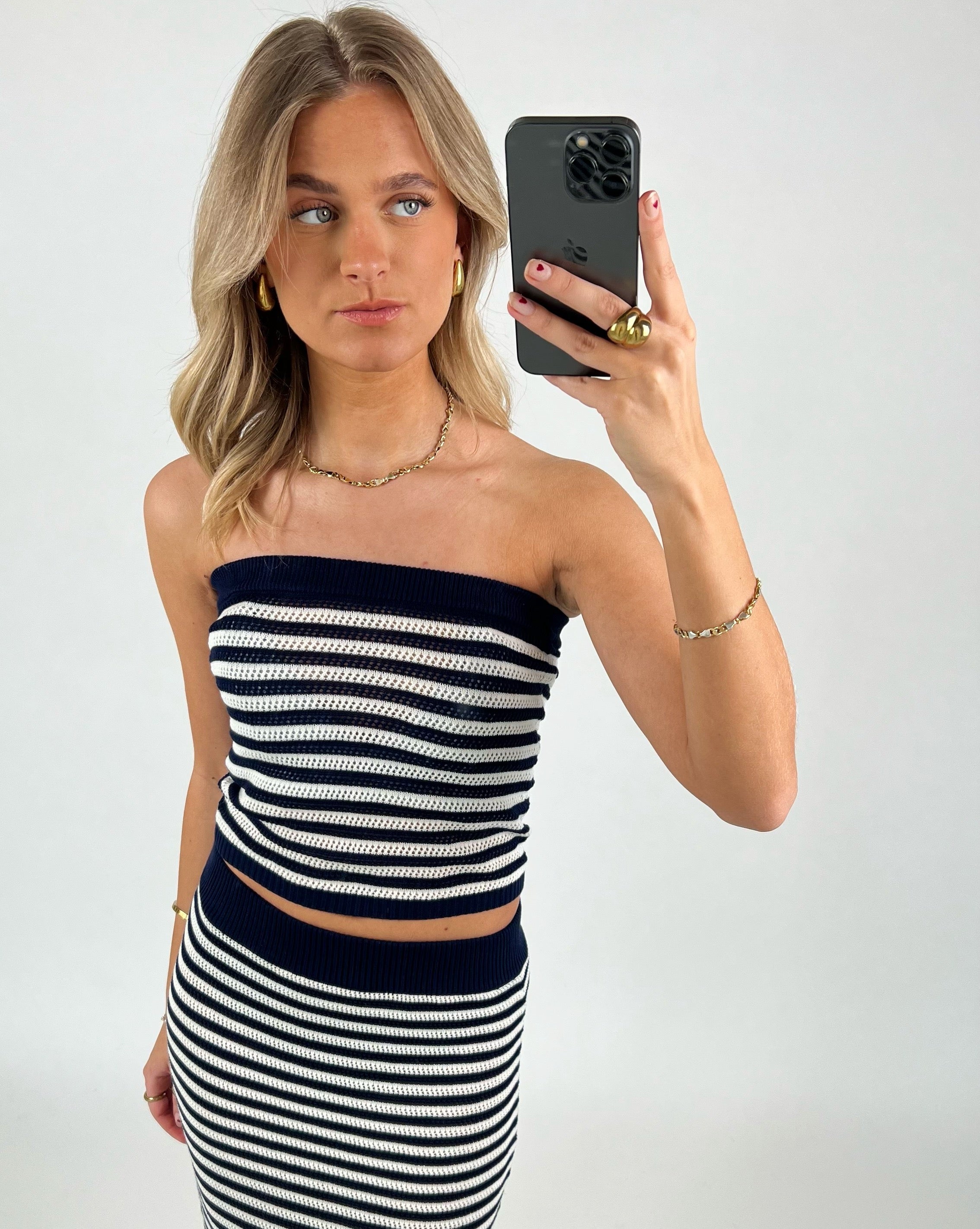 En kvinde iført en sort-hvid stribet kjole tager en selfie med en smartphone. Hun står i et neutralt rum, iført guldøreringe og smykker.