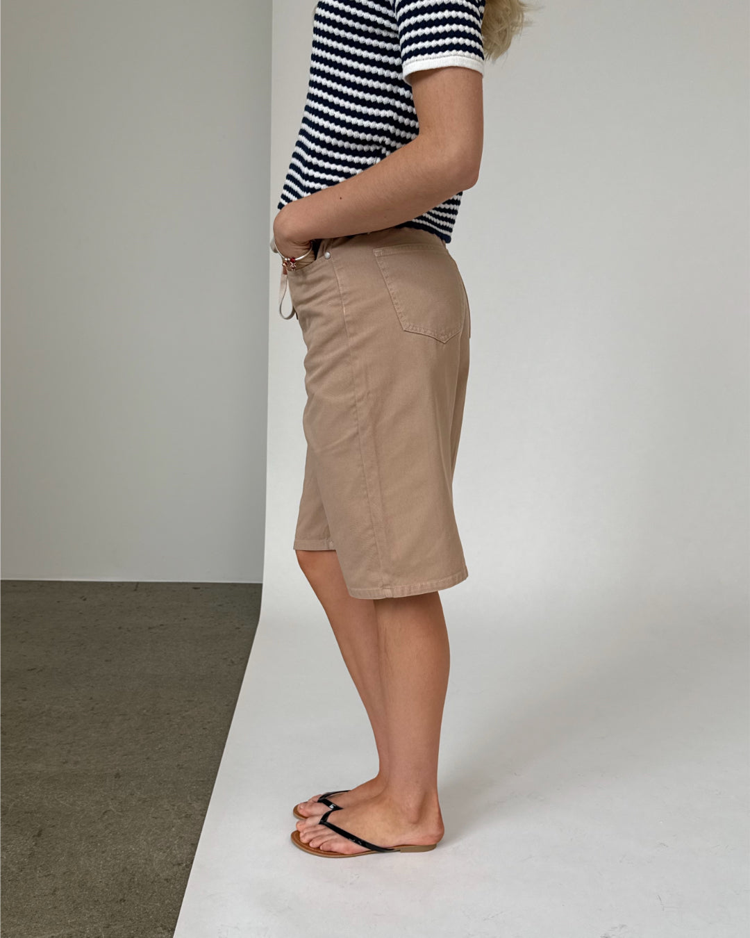 Person iført en stribet trøje og beige shorts står sidevendt, med hænderne i lommerne og iført sorte flip-flops. Baggrunden er enkel med en skillelinje mellem gulv og væg.