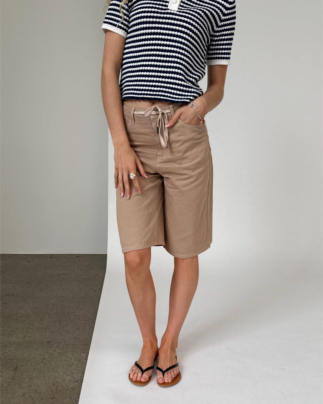 Person i stribet trøje og beige knælange shorts står oprejst med hænderne på siden. Flade sandaler bæres, og baggrunden er en todelt væg, grå og hvid.
