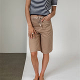 Person i stribet trøje og beige knælange shorts står oprejst med hænderne på siden. Flade sandaler bæres, og baggrunden er en todelt væg, grå og hvid.