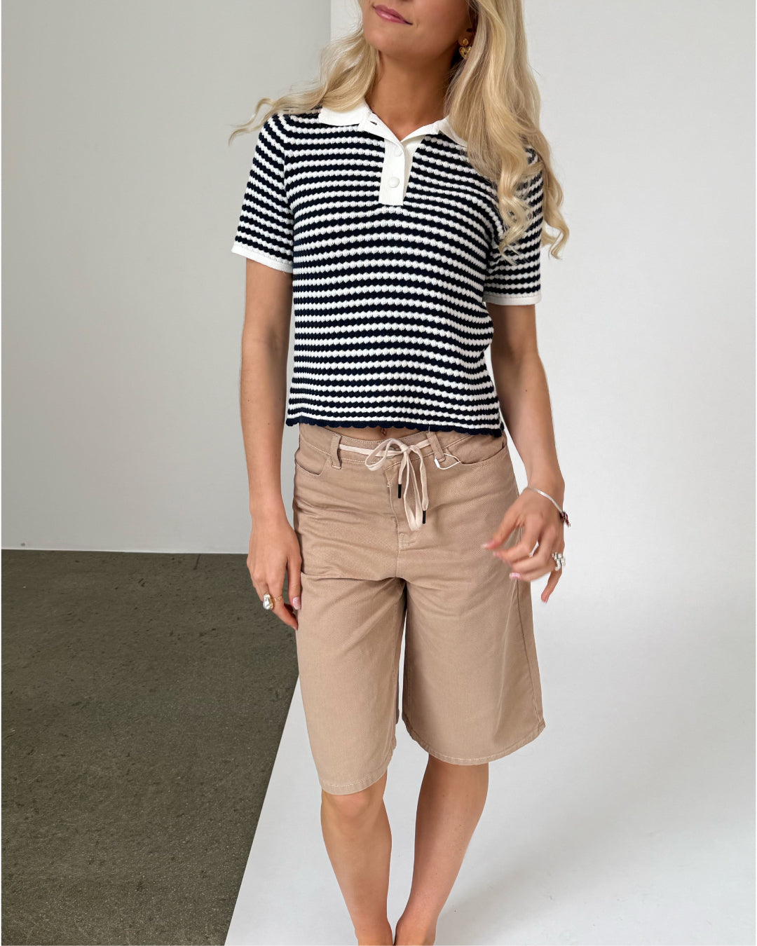 En person står iført en sort- og hvidstribet trøje og beige shorts. De står på et gulv med to forskellige farver, brun og hvid, i en enkel, lys tekstur.