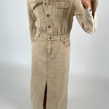 Lang, beige denimkjole med knapper, bliver båret af en person. Personen tager et spejlfoto i en minimalistisk, lys baggrundskontekst. Skoene har et skakternet mønster.