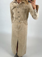 Lang, beige denimkjole med knapper, bliver båret af en person. Personen tager et spejlfoto i en minimalistisk, lys baggrundskontekst. Skoene har et skakternet mønster.