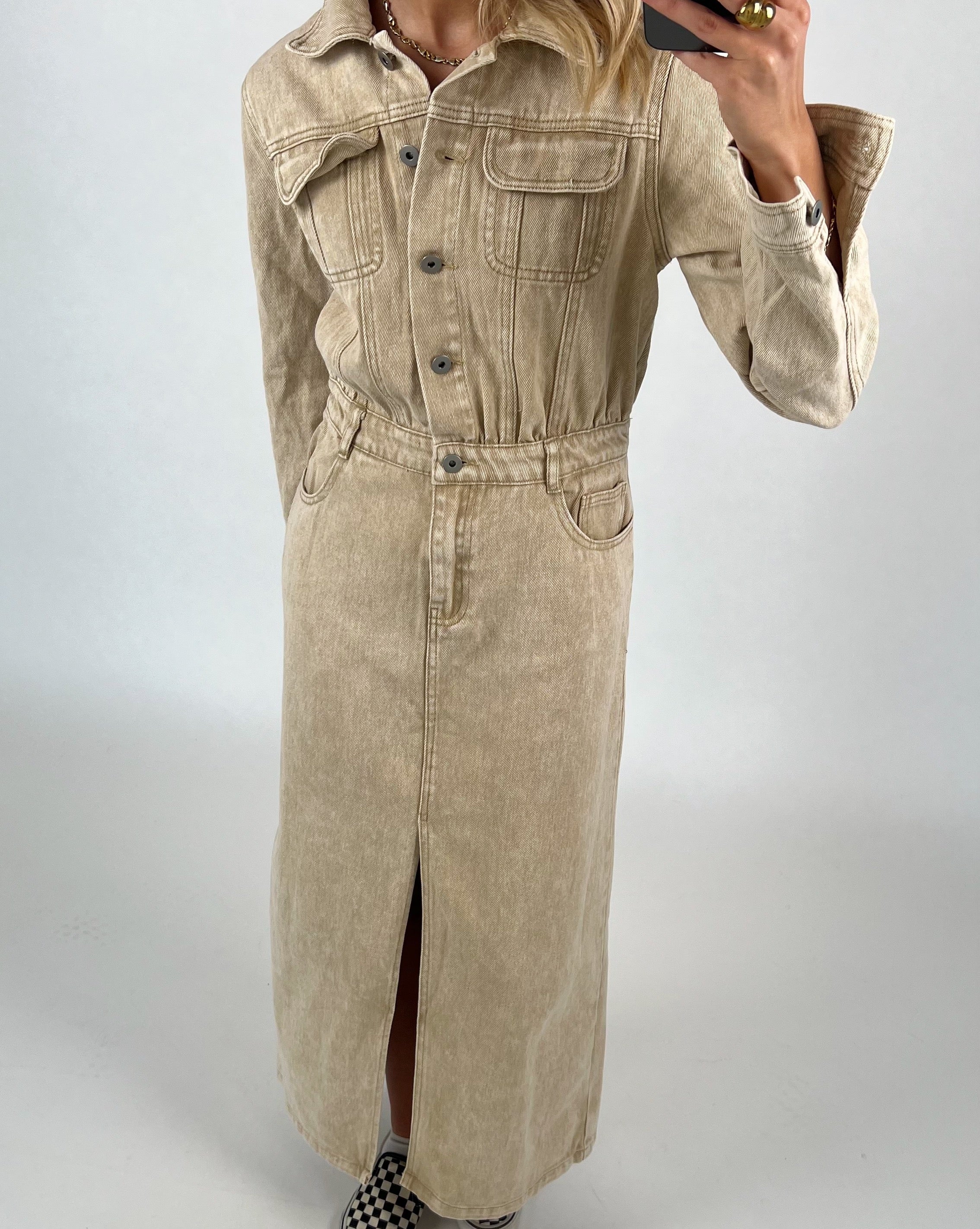 Lang, beige denimkjole med knapper, bliver båret af en person. Personen tager et spejlfoto i en minimalistisk, lys baggrundskontekst. Skoene har et skakternet mønster.
