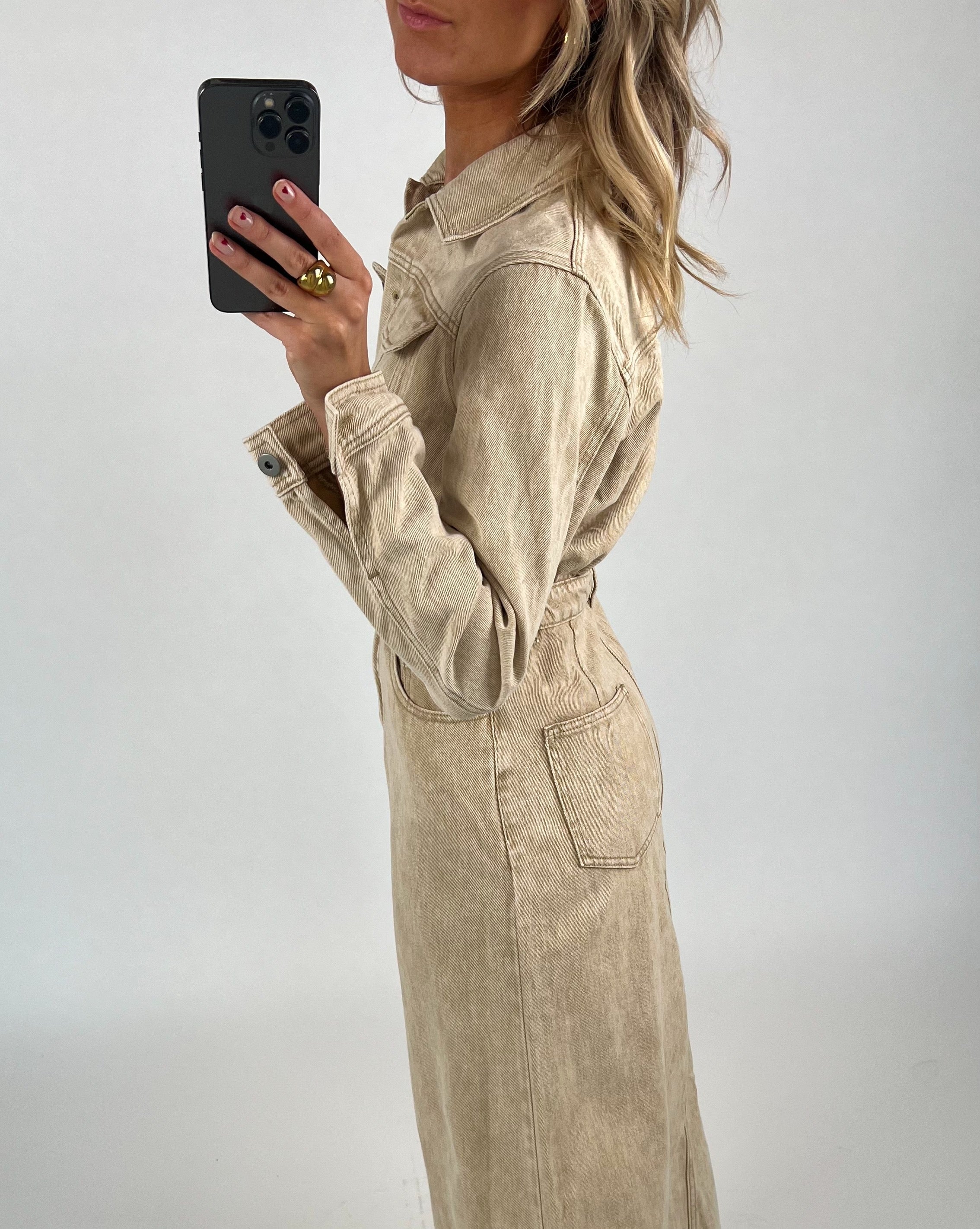 En person i en beige jakke holder en sort mobiltelefon med venstre hånd, mens de står mod en neutral, lysgrå baggrund.