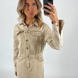 Person fotografere sig selv iført en beige denimkjole med knapper, poserer i en minimalistisk, lysegrå baggrund. Personen holder en sort smartphone og laver en sjov ansigtsudtryk.