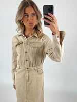 Person fotografere sig selv iført en beige denimkjole med knapper, poserer i en minimalistisk, lysegrå baggrund. Personen holder en sort smartphone og laver en sjov ansigtsudtryk.