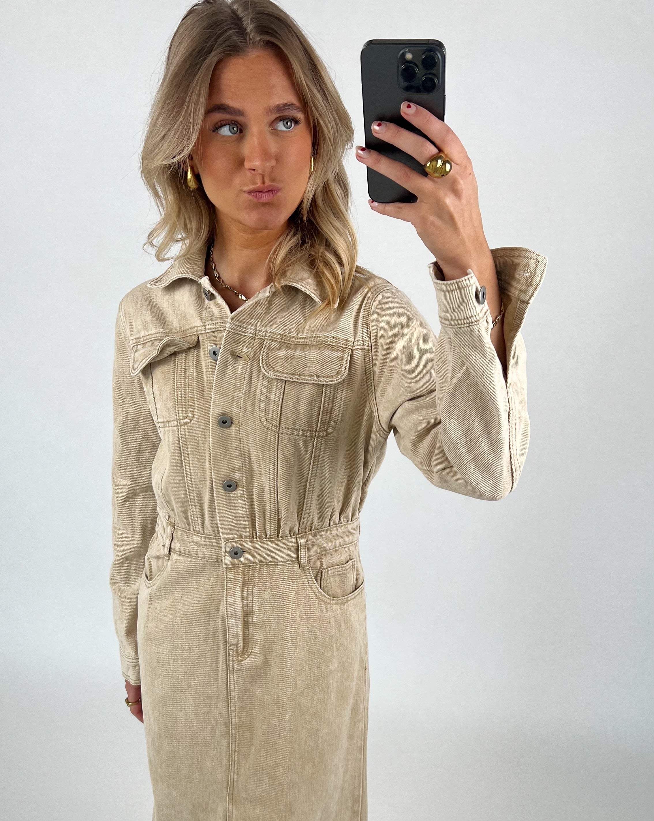 Person fotografere sig selv iført en beige denimkjole med knapper, poserer i en minimalistisk, lysegrå baggrund. Personen holder en sort smartphone og laver en sjov ansigtsudtryk.