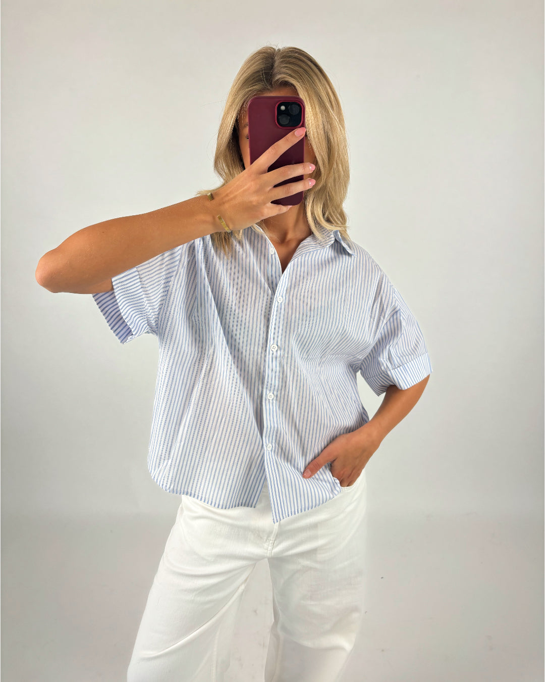 Person tager selfie med en rød telefon, iført en lyseblå stribet skjorte og hvide bukser, i et neutralt rum.
