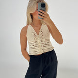En person tager en selfie med en mobiltelefon iført en cremefarvet, strikket top og sorte bukser i et minimalistisk, lyst rum.