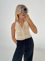 En person tager en selfie med en mobiltelefon iført en cremefarvet, strikket top og sorte bukser i et minimalistisk, lyst rum.