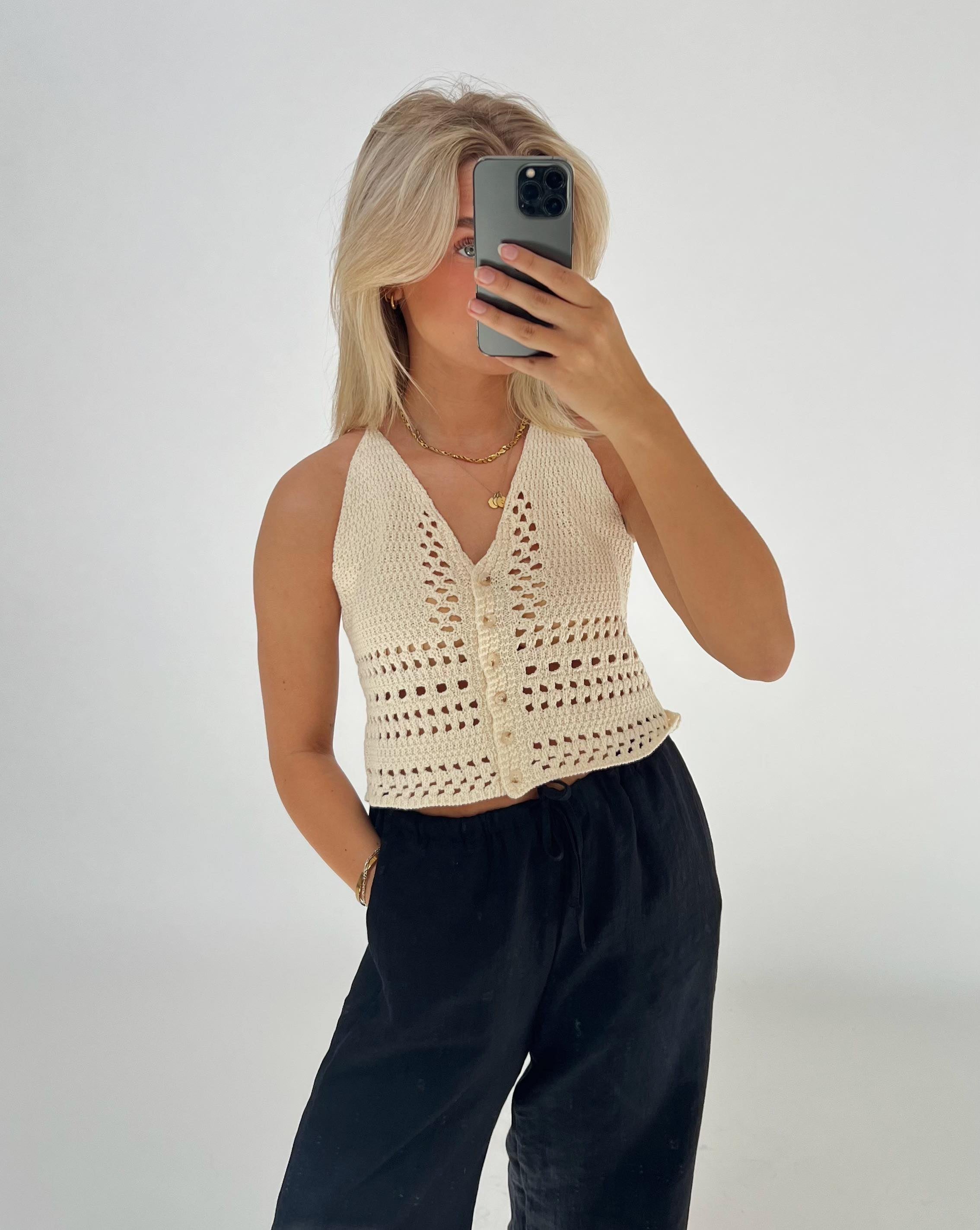 En person tager en selfie med en mobiltelefon iført en cremefarvet, strikket top og sorte bukser i et minimalistisk, lyst rum.