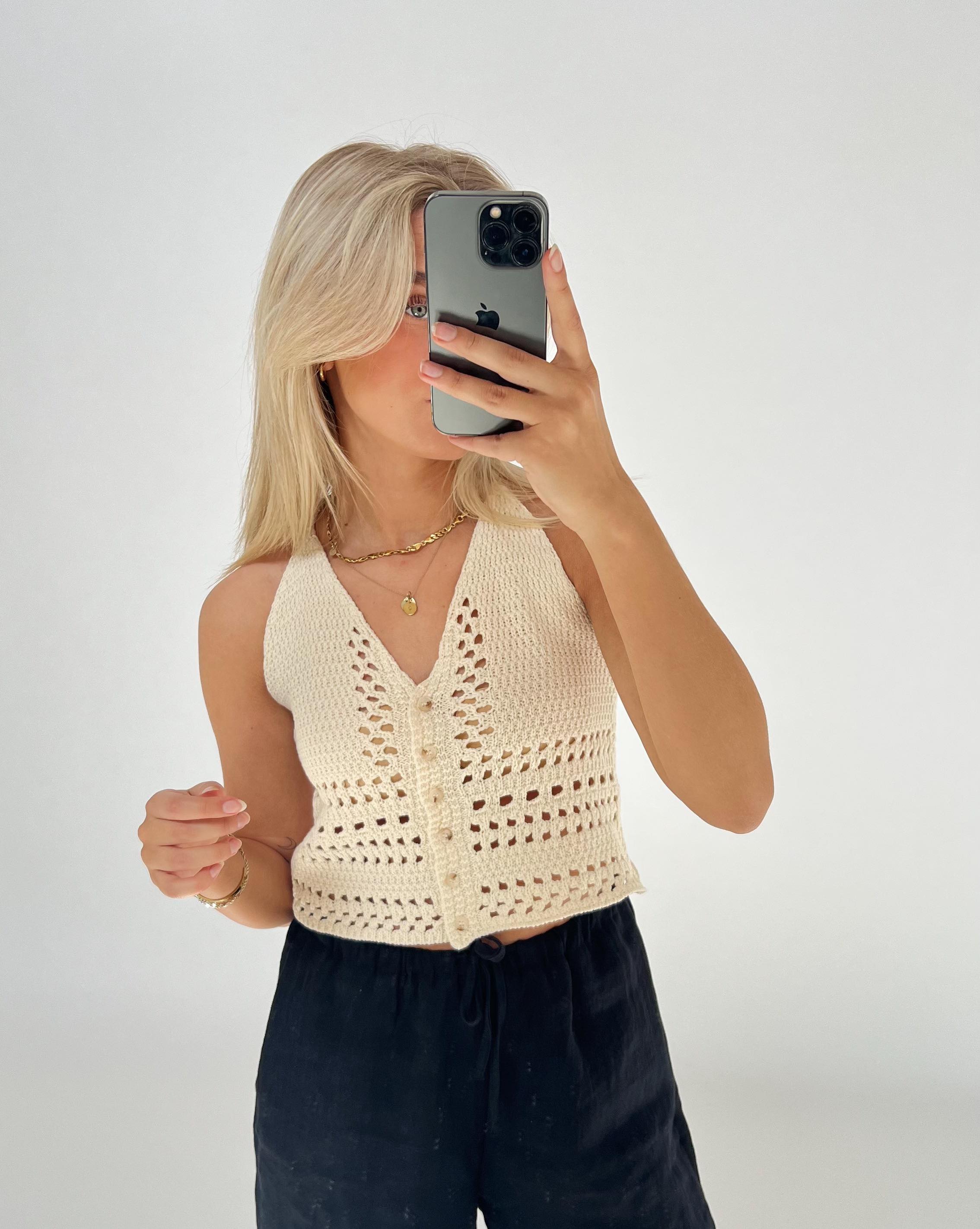 En person tager en selfie i et spejl, iført en cremefarvet strikket top og sorte bukser, med en smartphone i hånden, i et lyst rum.