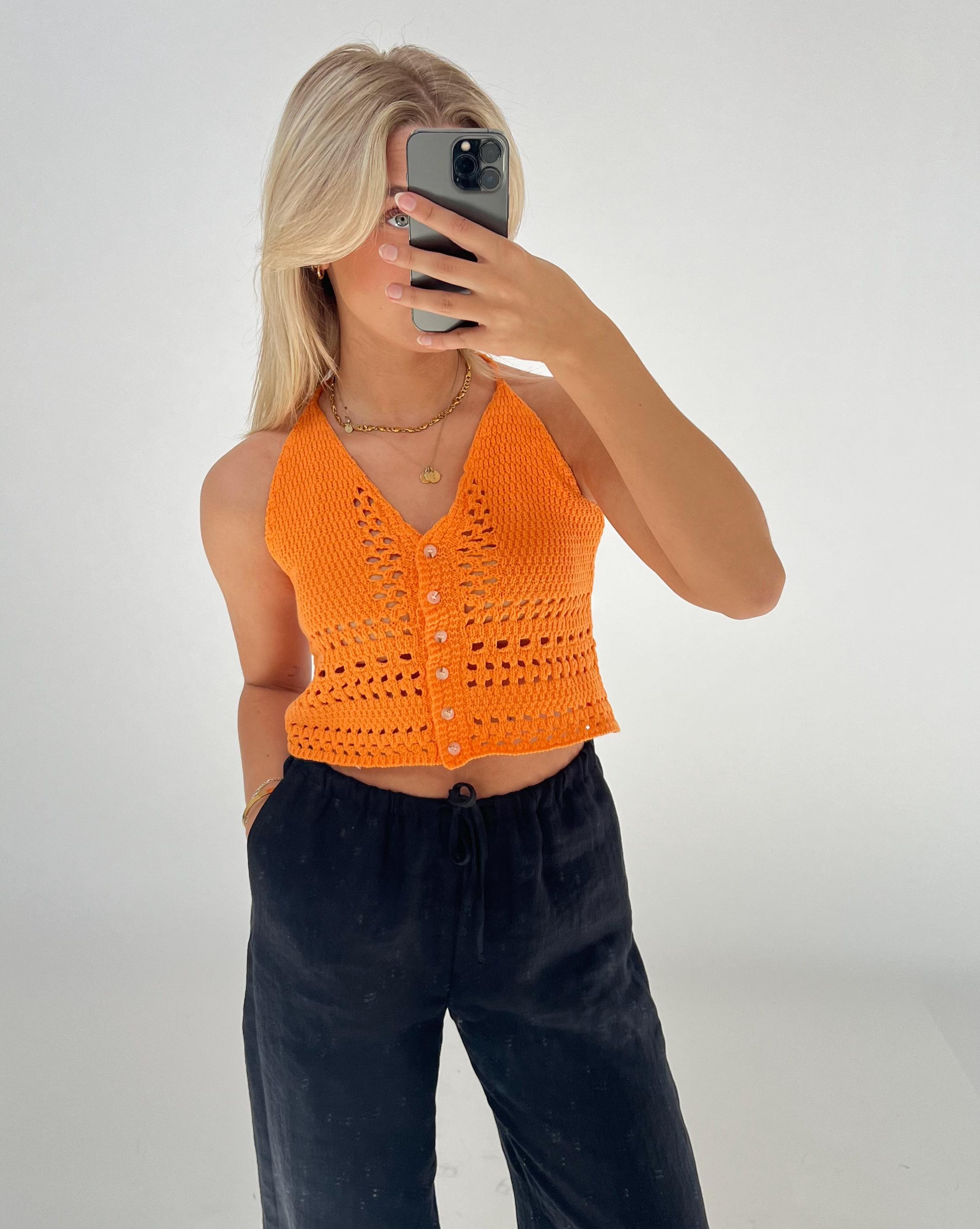 En person iført en orange strikketop fotograferer sig selv med en smartphone. Personen står i et rum med en neutral hvid baggrund og bærer sorte bukser.