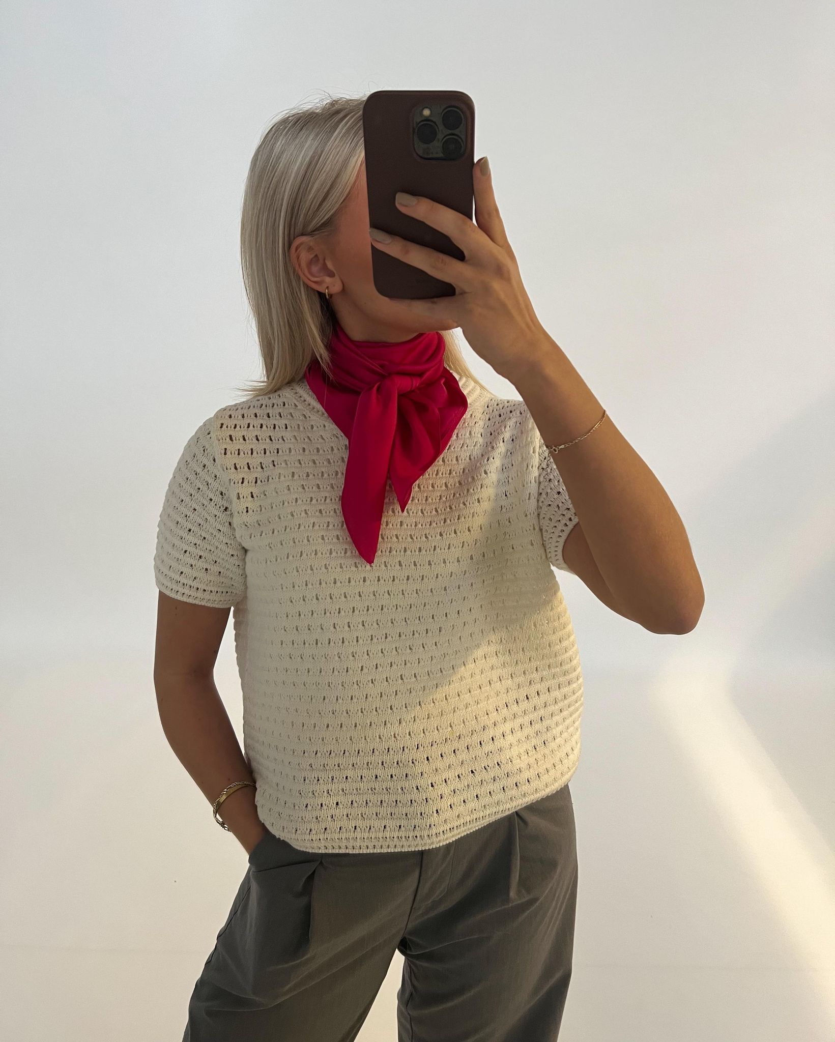 En person med langt, lyst hår tager et spejlbillede iført en hvid trøje og grå bukser. De har et rødt tørklæde om halsen. Baggrunden er neutral og ensfarvet.