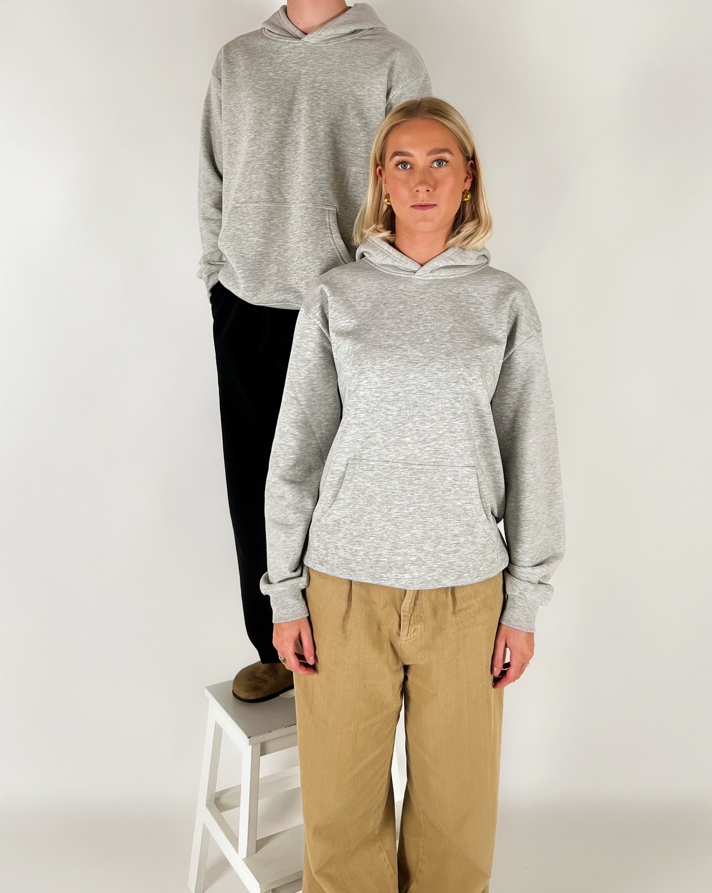 To personer iført grå hættetrøjer står, en på en skammel, mod en hvid baggrund; den ene bærer beige bukser.