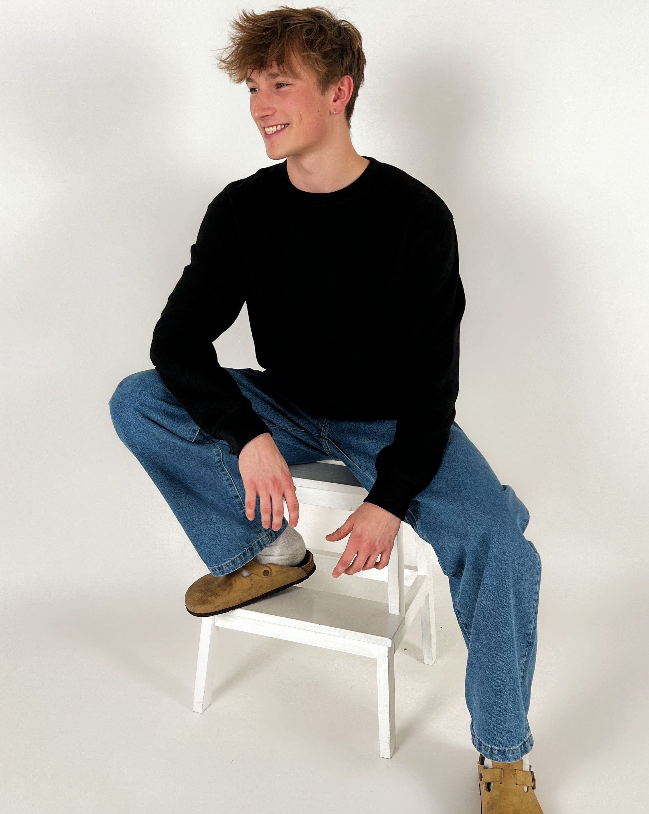 En ung mand sidder afslappet på en hvid skammel iført sort trøje, blå jeans og brune sko. Han smiler i et lyst studie.