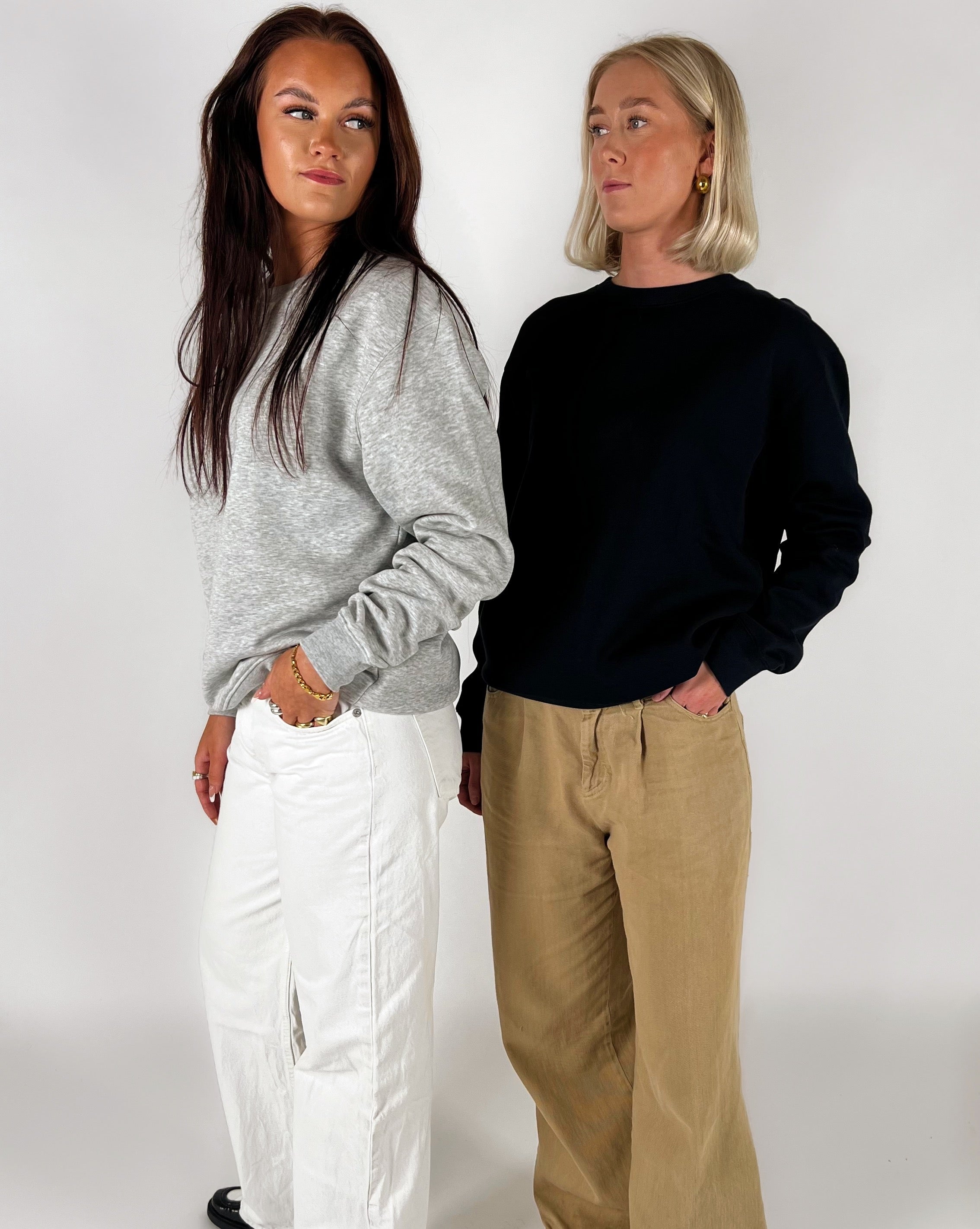 To kvinder står ved siden af hinanden. Den ene bærer en grå trøje og hvide bukser, mens den anden har en sort trøje og beige bukser på. Baggrunden er hvid.