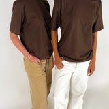 To personer iført brune t-shirts står i et neutralt rum. Den ene har beige bukser, den anden hvide. Begge står afslappet, delvist vendt mod hinanden.
