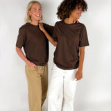 To personer i brune T-shirts smiler og står ved siden af hinanden; den ene har hvide bukser, den anden beige bukser, med en hvid baggrund.