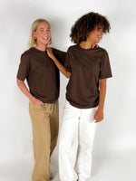 To personer i brune T-shirts smiler og står ved siden af hinanden; den ene har hvide bukser, den anden beige bukser, med en hvid baggrund.