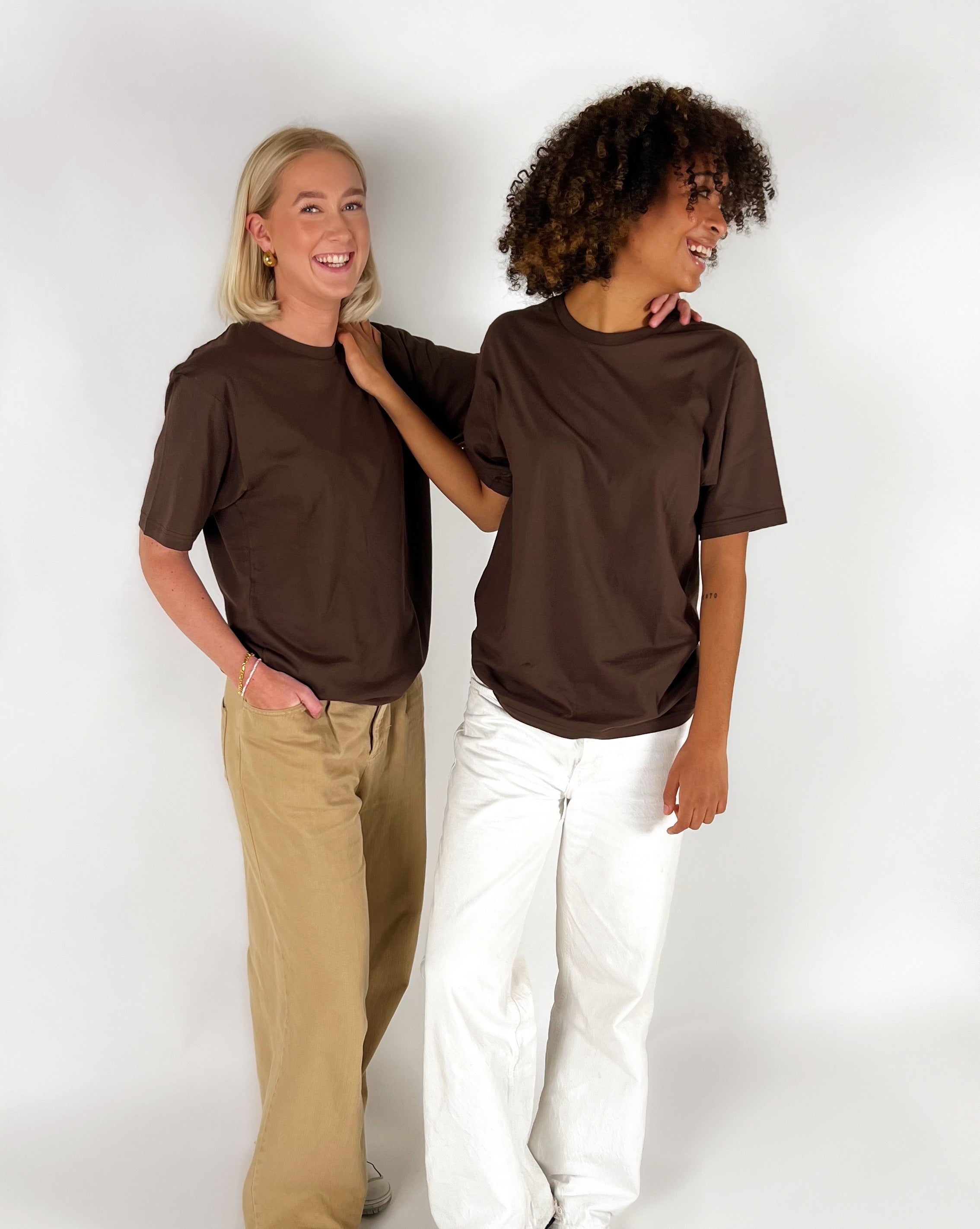 To personer i brune T-shirts smiler og står ved siden af hinanden; den ene har hvide bukser, den anden beige bukser, med en hvid baggrund.